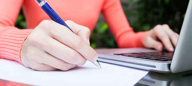 A woman, sitting in a garden, handwrites a resignation letter while viewing information on her laptop.