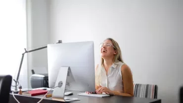 Persoon lacht achter groot computerscherm op bureau.