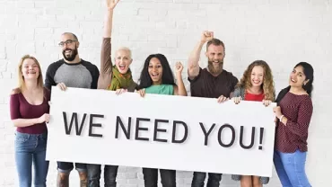 Groep mensen houdt een bord vast met de tekst 'WE NEED YOU!'