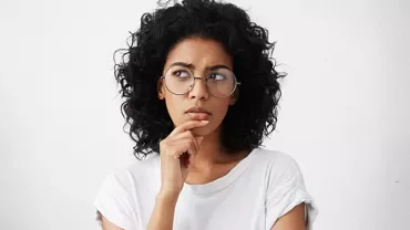 Person with curly hair and glasses looks thoughtful.