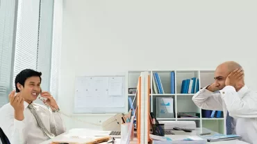 Two people in an office, one is on the phone, the other covers their ears.