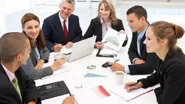 Business meeting with six people around a table with documents and a laptop.