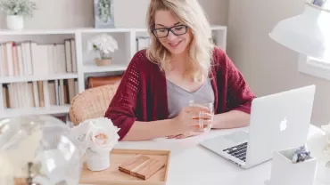 Persoon met bril drinkt thee bij laptop aan bureautafel.