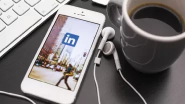 iPhone met LinkedIn-app naast kopje koffie en oordopjes op donker bureau.