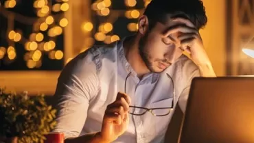 Person at desk with glasses in hand, looking at a laptop, under warm lighting