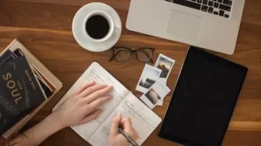 Een persoon schrijft in een notitieboek op een bureau met laptop, koffie en foto's.