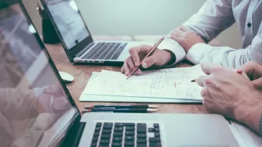Twee personen werken aan een bureau met laptops en papier.