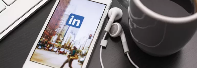 Smartphone displaying LinkedIn app beside a cup of coffee and earphones on a desk.