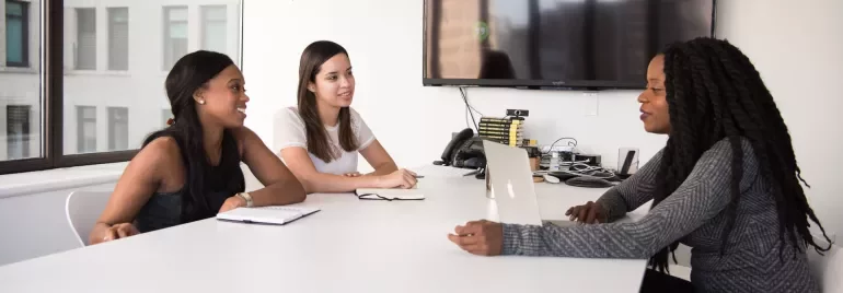 Three people in a meeting room with a laptop and a TV in the background.
