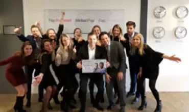 Een groep mensen poseert lachend met een bord in een kantoor.