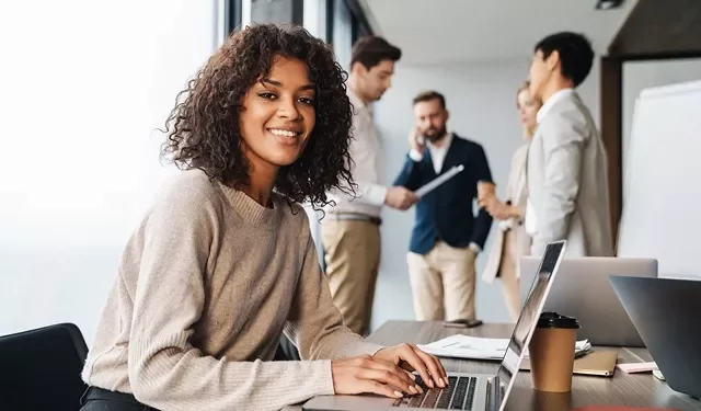 Person with curly hair using a laptop, colleagues in background.